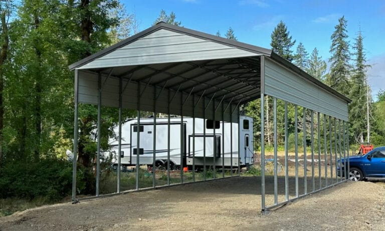 Large metal carport beside trailer and truck in forest.